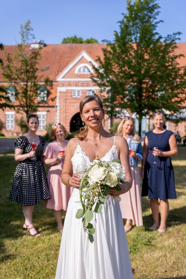 Eine lächelnde Braut im weißen Kleid hält einen Blumenstrauß und steht mit vier Frauen in farbenfrohen Kleidern hinter ihr im Freien - ein Beispiel für authentische Hochzeitsbilder von einem Hochzeitsfotograf aus Lübeck, vor einem Backsteingebäude und grünen Bäumen.
