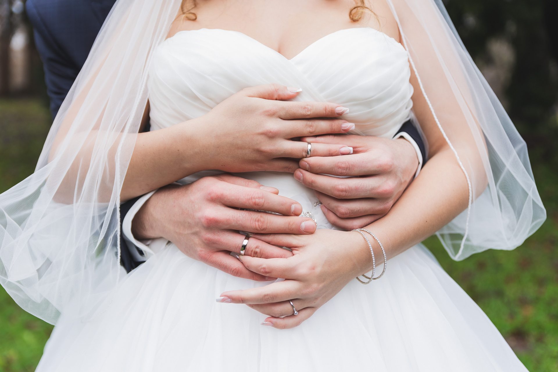 Eine Nahaufnahme von Braut und Bräutigam, die sich umarmen, wobei sich ihre Hände sanft um ihre Taille legen. Das weiße trägerlose Kleid der Braut schimmert, während ihre Eheringe das Licht einfangen - ein Moment natürlicher Hochzeitsfotografie vor verschwommenem, grünem Hintergrund.